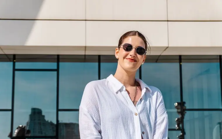 Woman in sunglasses and a white shirt standing in front of a glass building on a sunny day