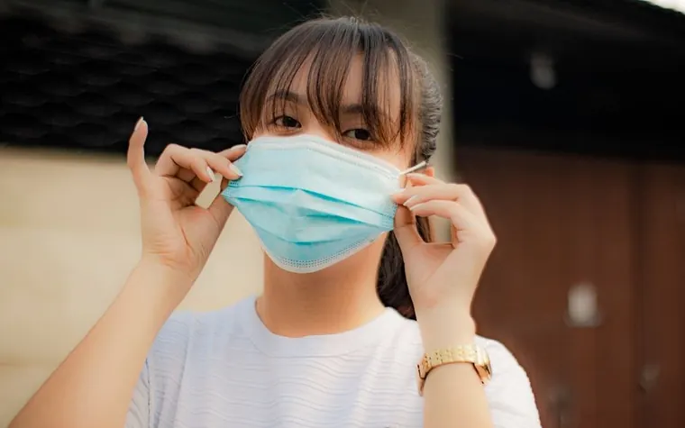 Young woman with bangs pulling a light blue surgical mask over her face outdoors