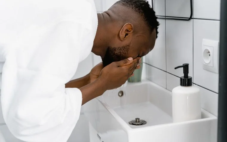 Man in a white shirt leaning over a bathroom sink to wash his face with water