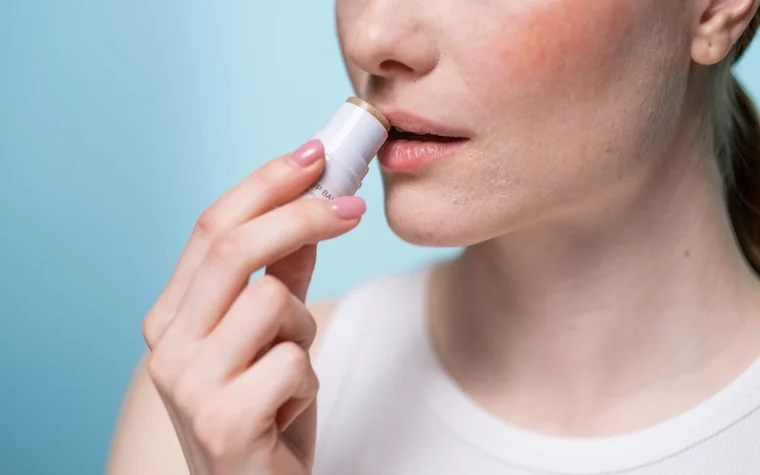 Woman holding a white lip balm stick against her lips in front of a pale blue wall