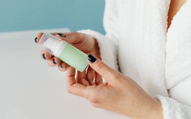 Person in a bathrobe holding a green pump bottle of skincare product