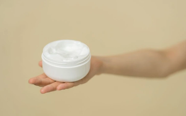 A hand holding an open jar of white moisturiser