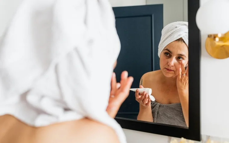 A woman applying face cream in front of a bathroom mirror