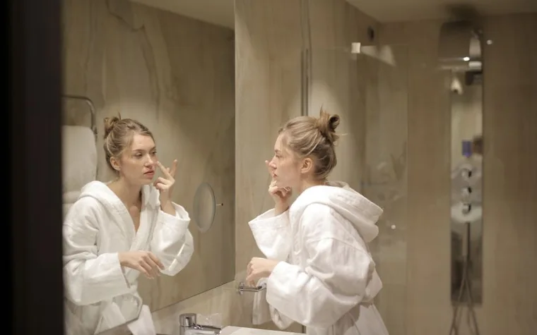 A woman in a bathrobe applying moisturizer while looking in a mirror
