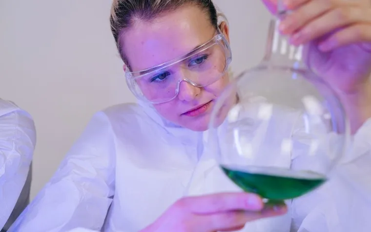 A researcher in safety goggles examining a green liquid in a flask