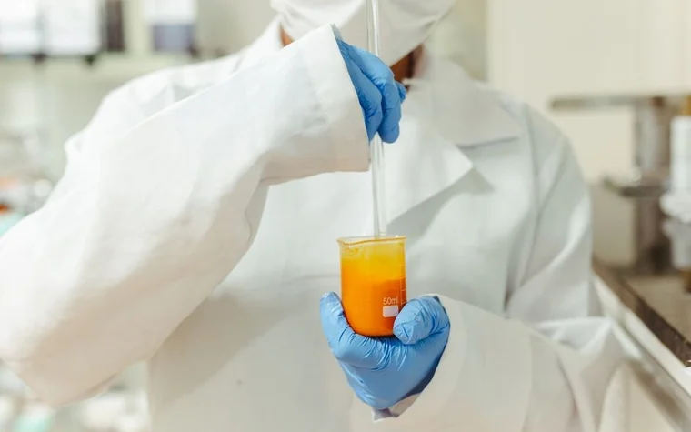 A gloved hand using a dropper over an orange liquid in a beaker