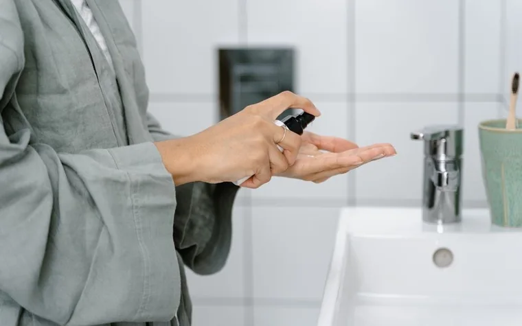 A hand applying a skincare product at a bathroom sink, soft morning light, bathrobe sleeve visible