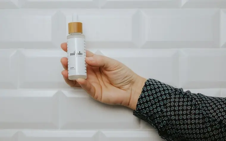 Close-up macro shot of a glass dropper bottle held above skin, a single amber-toned serum droplet suspended at the tip catching soft light, minimal clean background, no text or logos visible