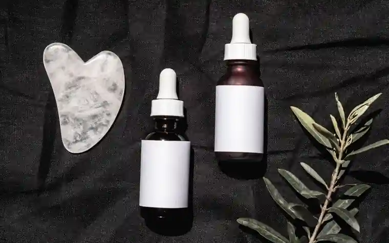 A gua sha stone and dropper bottles resting on dark fabric with a green leaf