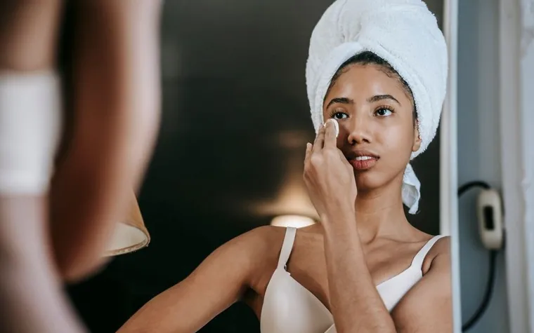 Woman with a hair towel applying skincare near a mirror