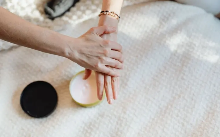 Woman's hands smoothing moisturizer over the back of one hand on a cream blanket
