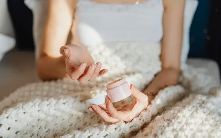 A woman seated under a knit blanket scooping cream from a small jar