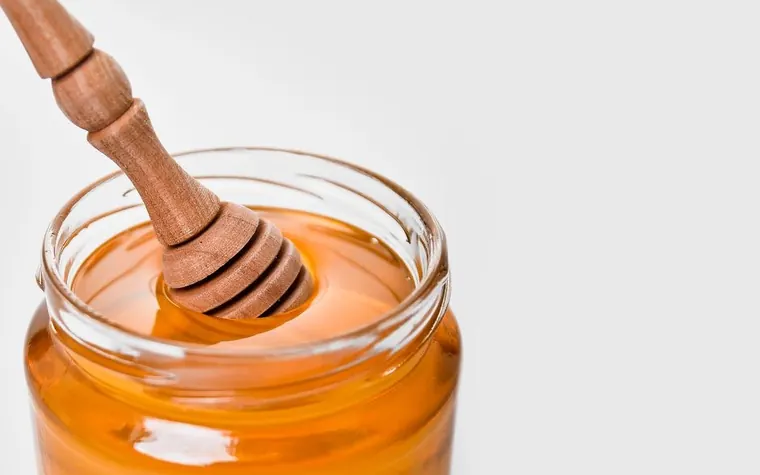 Glass jar of golden honey with a wooden dipper resting in it against a white background