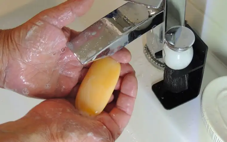 Hands covered in foamy soap lather holding a yellow bar of soap under a running tap