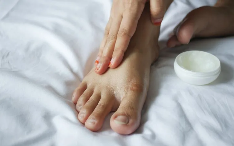 Close-up of a person rubbing moisturizer into the top of a bare foot on white bedding