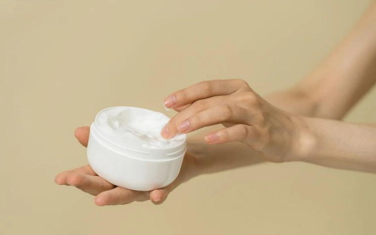 A hand holding an open jar of white cream while the other scoops some out