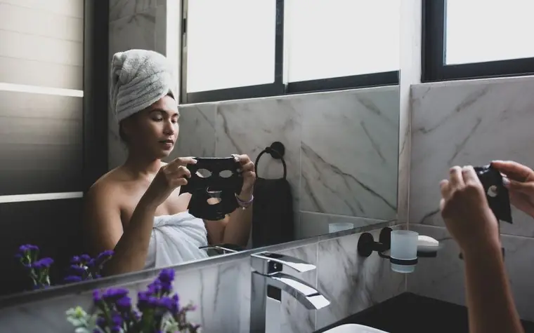 A woman applying a hydrating sheet mask while looking in a bathroom mirror