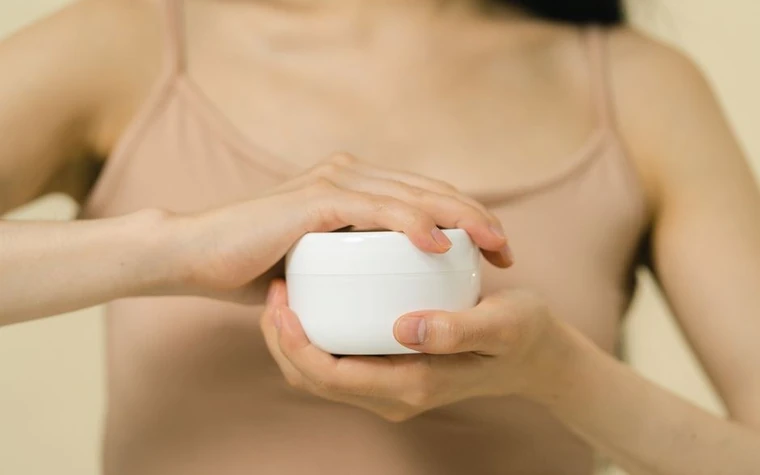 A woman holding a round jar of moisturizer close to her collarbone