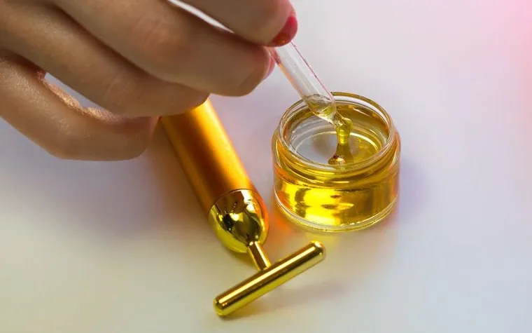 A hand holding a gold dropper above a jar of golden serum