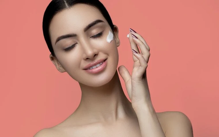 Woman applying a dab of cream to her cheek against a coral background