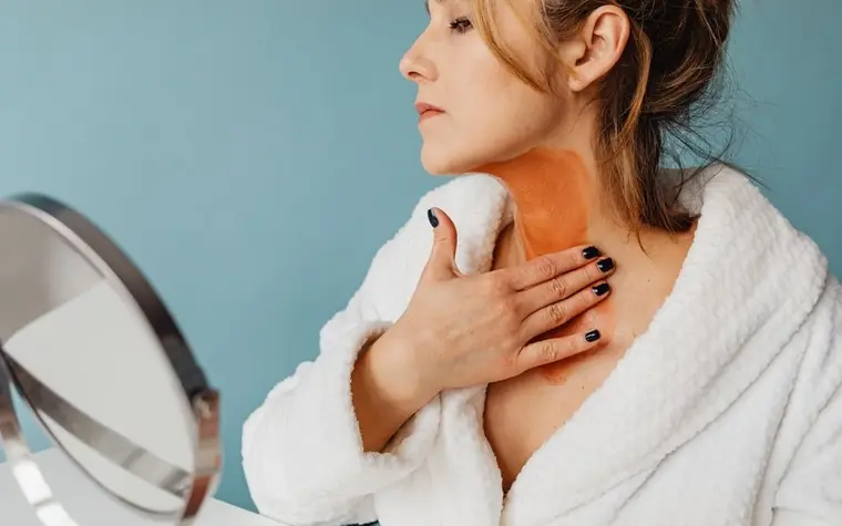 Woman in a white bathrobe applying a clay mask to her neck in a bright bathroom
