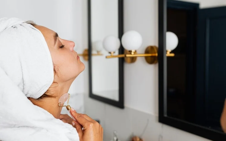 Woman with a towel wrapped around her hair holding a skincare dropper bottle near a bathroom mirror