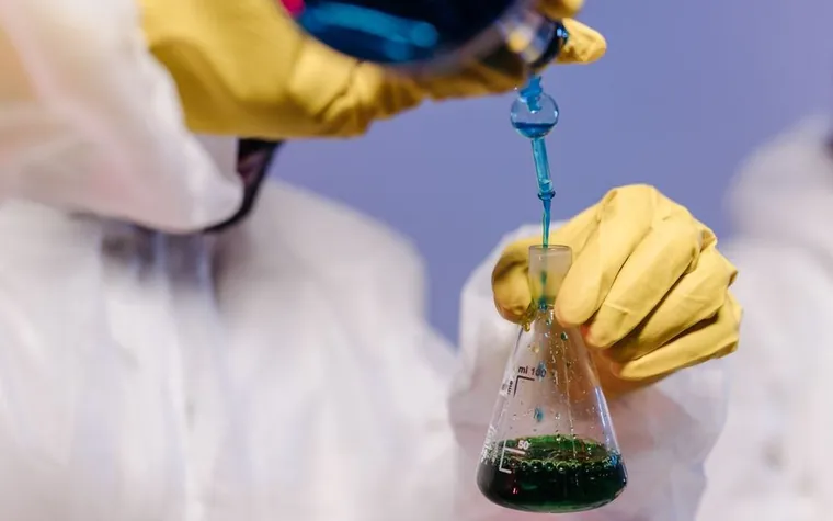 Gloved hands mixing a blue solution into a green liquid in a laboratory flask