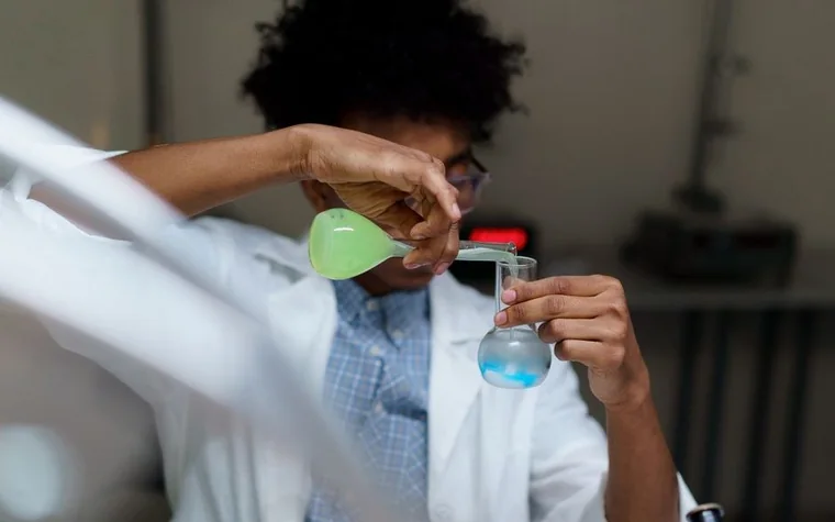A researcher in a lab coat pouring a green solution into a flask of blue liquid