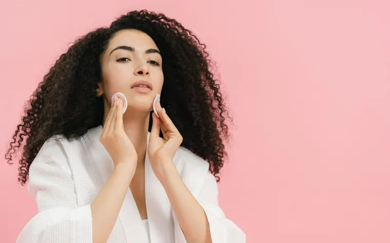 Young woman with curly hair pressing a cotton pad to her cheek to remove makeup