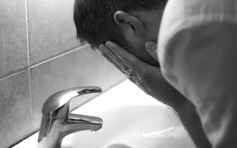 Black-and-white photo of a person splashing water on their face over a bathroom sink