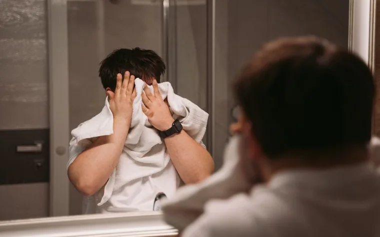 Man standing at a bathroom mirror wiping his face with a towel