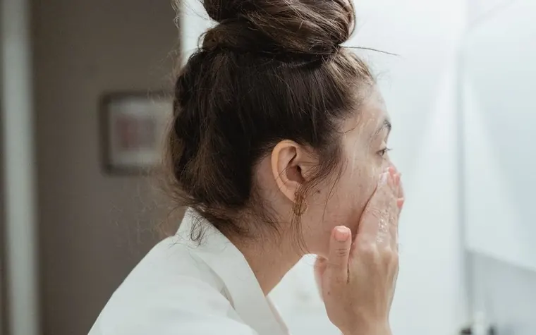 A woman in a bathrobe checking her skin in a bathroom mirror