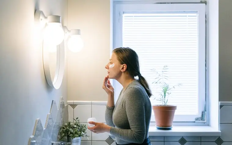 A woman applying face cream in a bright bathroom