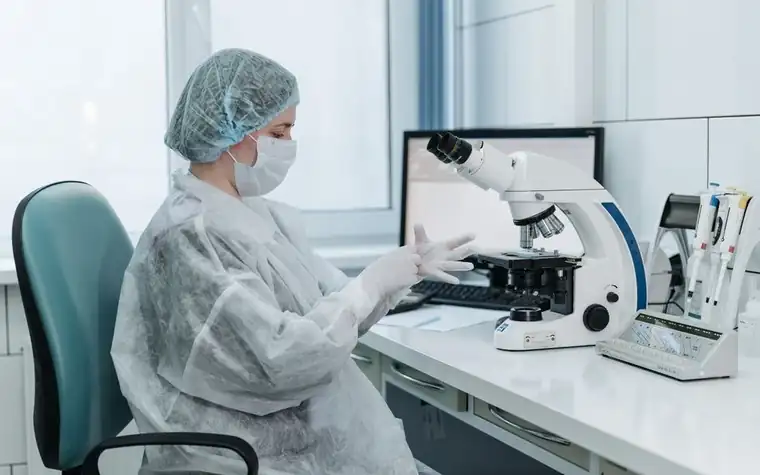 Lab technician in protective gear and mask using a microscope at a clinical workstation