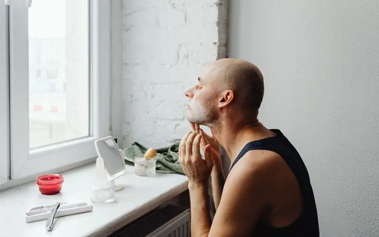 Man applying a skincare product to his face while looking out a window