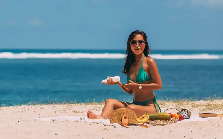 Woman in sunglasses sitting on a sandy beach squeezing sunscreen into her hand beside a straw bag