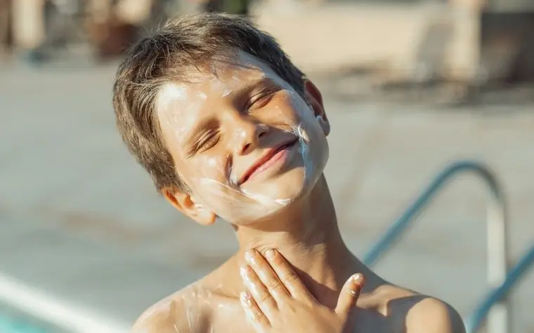 Smiling boy with eyes closed rubbing white sunscreen across his face beside a pool