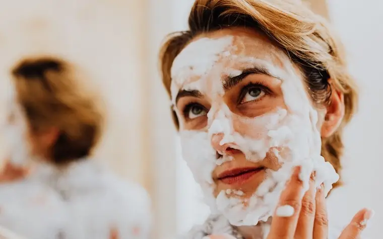 Woman with foaming cleanser on her face looking up while rinsing in a bright bathroom