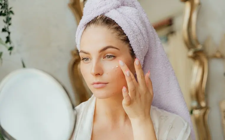 A woman with a hair towel applying moisturiser in front of a mirror