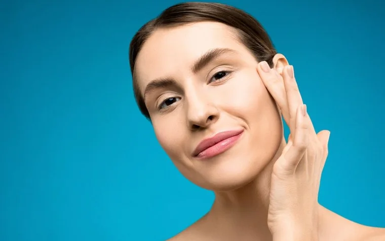 A woman touching her cheek with her fingertips against a plain background