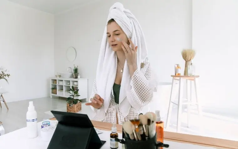 A woman in a bathrobe applying a skincare product in front of a mirror