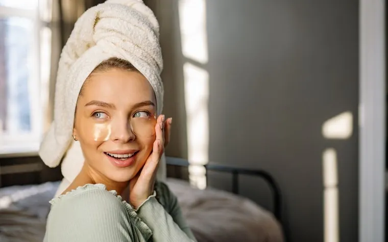 A woman with a hair towel applying cream to her face in bright light