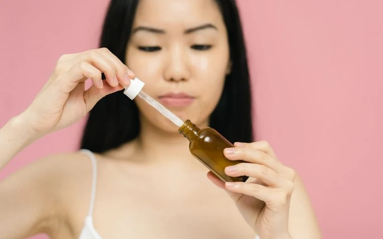 Woman holding an amber dropper bottle of serum against a pink background