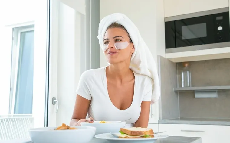 Woman wearing under-eye patches while having breakfast in a bright kitchen