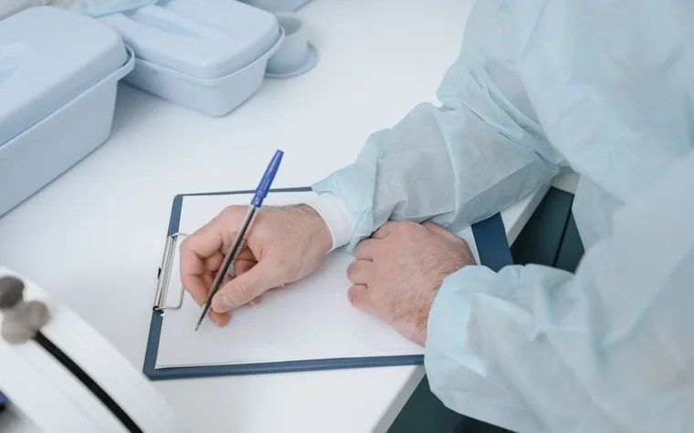 Gloved hand in a lab coat writing notes on a clipboard beside laboratory equipment