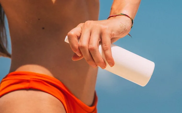 Woman applying sunscreen from a white bottle onto her arm against a blue sky