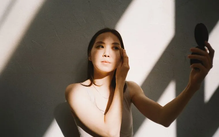 Woman holding a small mirror in a patch of sunlight and shadow on a wall