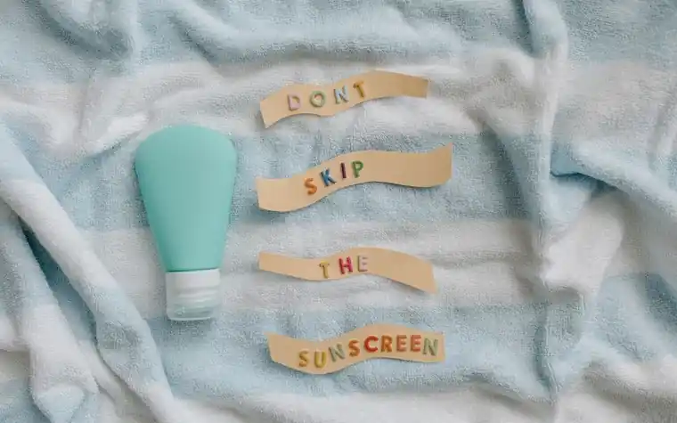 Flat lay of a teal sunscreen tube on a blue towel with paper letters reading don't skip the sunscreen