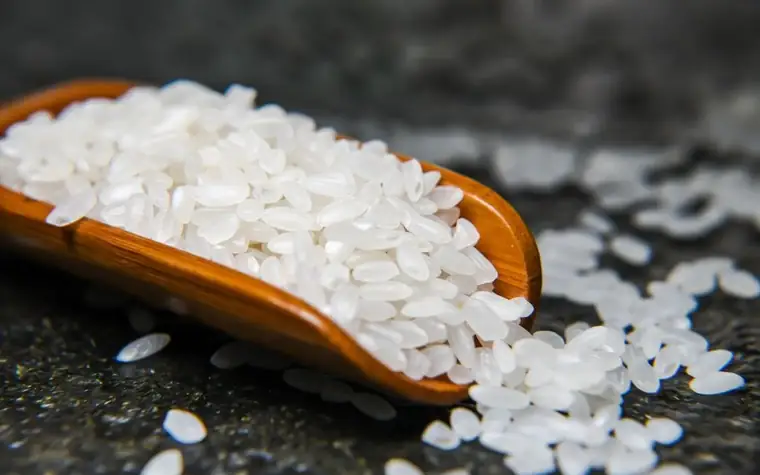 White rice grains piled on a wooden scoop on a dark surface