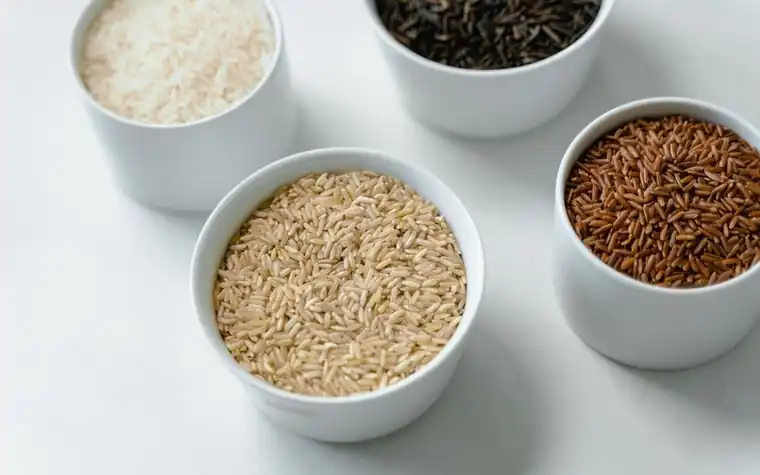 Four white ceramic bowls holding different types of rice grains on a pale table
